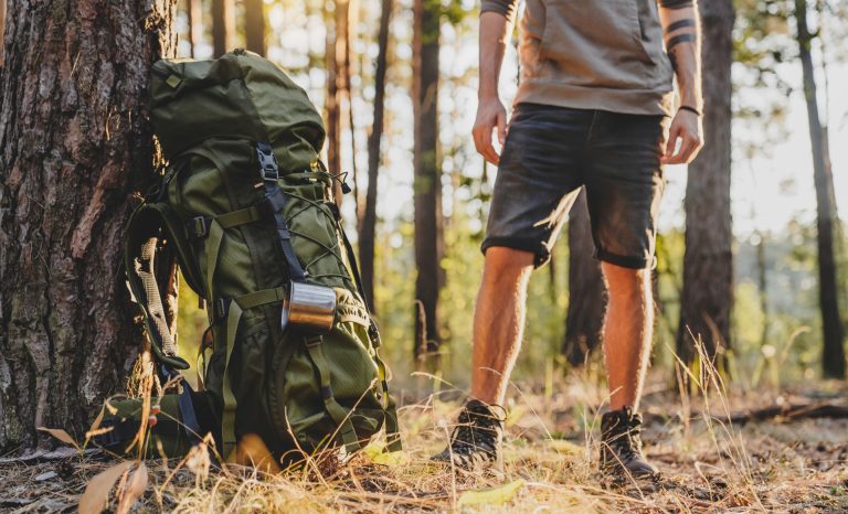 Cropped image of male hiker standing near his camping backpack in forest. Away from everything concept. Adventurer explorer traveler in woods in summer